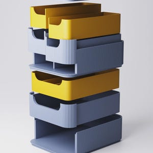 Op de afbeelding: Een stapelbare opbergorganizer met een deksel. De organizer heeft afwisselend gele en blauwgrijze lades. De lades hebben een geribbeld ontwerp aan de voorkant en een uitsparing voor een gebogen handgreep. Het deksel en de basis zijn ook blauwgrijs.
