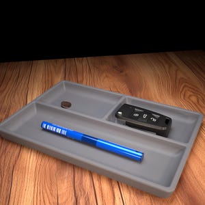 May include: A gray rectangular tray with three compartments. A blue pen, a car key, and a coin are inside the tray. The tray is on a wooden surface.