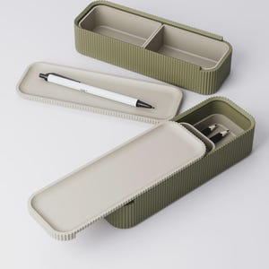 Peut inclure: Trousse &agrave; crayons rectangulaire vert olive avec une texture c&ocirc;tel&eacute;e et un couvercle gris clair. Le bo&icirc;tier est ouvert, r&eacute;v&eacute;lant un int&eacute;rieur divis&eacute;. Un stylo blanc &agrave; pointe noire repose sur le couvercle. Deux crayons sont &agrave; l'int&eacute;rieur.