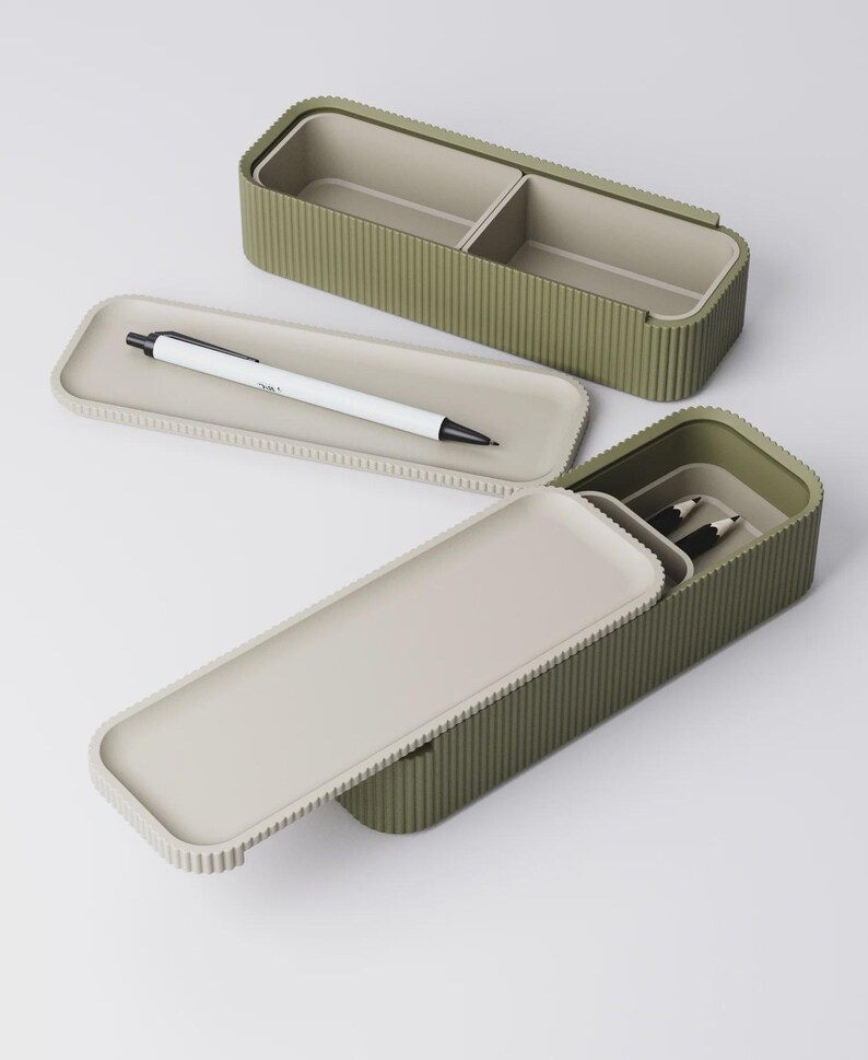 Peut inclure: Trousse &agrave; crayons rectangulaire vert olive avec une texture c&ocirc;tel&eacute;e et un couvercle gris clair. Le bo&icirc;tier est ouvert, r&eacute;v&eacute;lant un int&eacute;rieur divis&eacute;. Un stylo blanc &agrave; pointe noire repose sur le couvercle. Deux crayons sont &agrave; l'int&eacute;rieur.