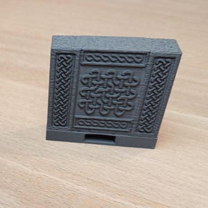 Op de afbeelding: Een grijze 3D-geprinte doos met een Keltisch knoopontwerp aan de voorkant. De doos heeft een kleine opening aan de onderkant.