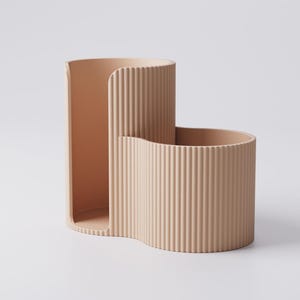 Puede incluir: Un organizador de escritorio beige moderno con un dise&ntilde;o ondulado &uacute;nico. El organizador tiene una secci&oacute;n alta y abierta y una secci&oacute;n m&aacute;s corta y curva, ambas con detalles acanalados verticales. El color neutro y el estilo minimalista son adecuados para cualquier decoraci&oacute;n.