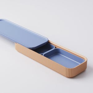 Puede incluir: Una fiambrera rectangular con exterior acanalado marr&oacute;n claro e interior azul claro. La tapa est&aacute; abierta, revelando dos compartimentos azules. La fiambrera tiene un dise&ntilde;o moderno.