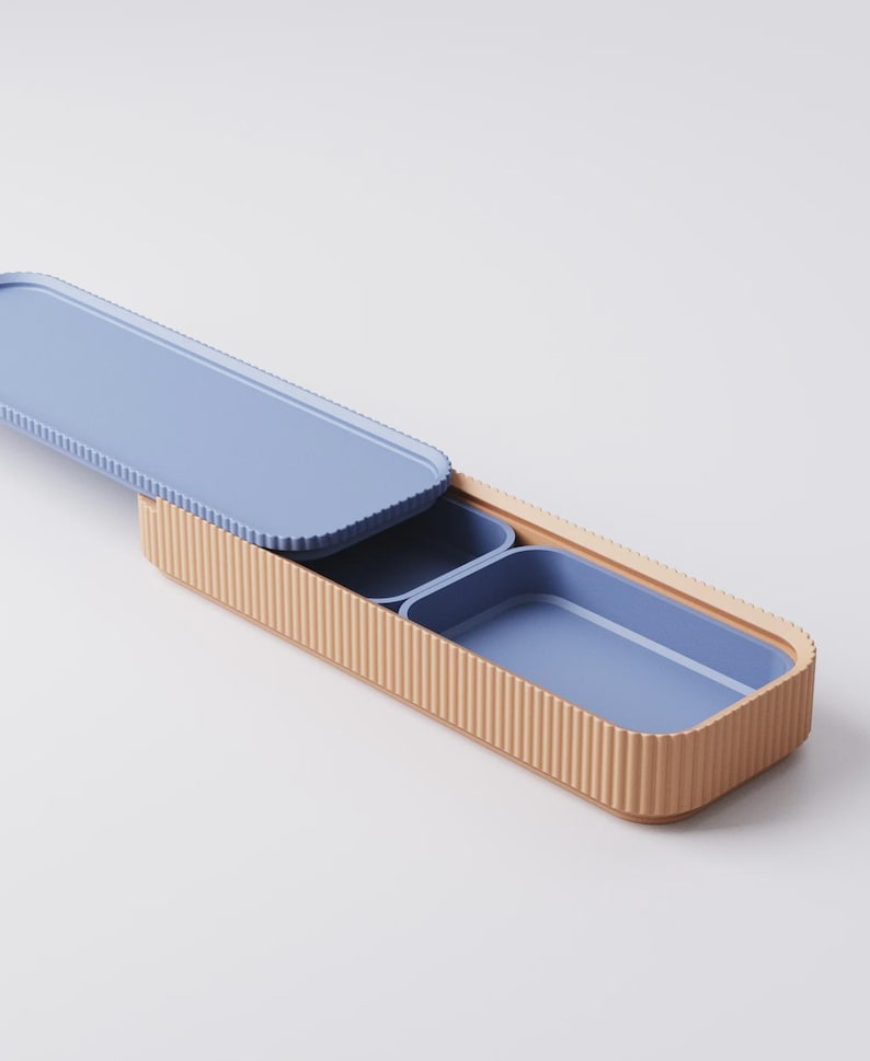 Puede incluir: Una fiambrera rectangular con exterior acanalado marr&oacute;n claro e interior azul claro. La tapa est&aacute; abierta, revelando dos compartimentos azules. La fiambrera tiene un dise&ntilde;o moderno.