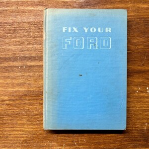 May include: A light blue hardcover book titled FIX YOUR FORD in white lettering on the front cover. The book has a rectangular shape with a visible spine on the left side and shows minor signs of wear on the surface of the blue fabric cover.