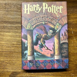 May include: A hardcover book titled Harry Potter and the Sorcerer's Stone by J.K. Rowling. The front cover features a colorful illustration of a character flying on a broomstick through a stone archway, with a unicorn and castle in the background.