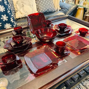 Puede incluir: Una colección de vajilla de vidrio rojo rubí, que incluye platos cuadrados, cuencos y tazas de té con platillos. El conjunto está dispuesto sobre una superficie reflectante, mostrando el rico color y las formas geométricas de las piezas.