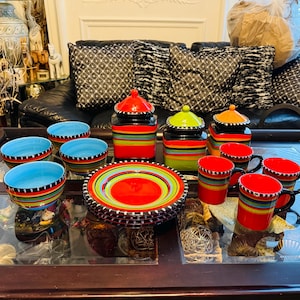 Puede incluir: Un colorido juego de vajilla de cerámica con cuencos, platos y tazas. El juego incluye cuencos con interior azul claro y platos con centro rojo. Las piezas están decoradas con rayas horizontales de varios colores.