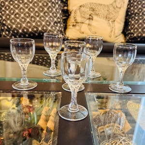 Op de afbeelding: Zes heldere wijnglazen met een geribbeld ontwerp op de kom en een delicate, getextureerde steel. De glazen zijn gerangschikt op een reflecterend oppervlak, dat hun elegante vorm en helderheid laat zien. De achtergrond omvat decoratieve kussens.
