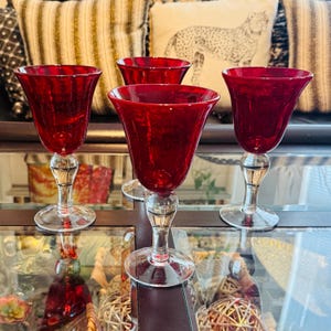 May include: Four ruby red wine glasses with clear stems and bases. The glasses have a tulip-shaped bowl and a decorative stem. The glasses are arranged on a mirrored surface, with decorative pillows in the background.