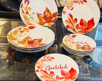 May include: A collection of white ceramic plates and bowls featuring a vibrant autumn leaf pattern in shades of red, orange, and yellow. Some pieces display the word Grateful in a red script font, while others show only the colourful foliage design.