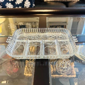May include: A clear glass serving tray with four divided sections. The tray has a raised rim and a decorative design of grapes and leaves.