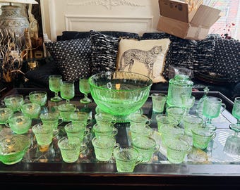 May include: A collection of vintage green glassware, including cups, bowls, and serving dishes, arranged on a dark table. The glassware features a floral pattern. A large glass bowl is in the center. The set is displayed in a well-lit room.