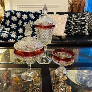May include: Three clear glass candy dishes with a red band around the top. The dishes are on a glass table with a decorative pattern.
