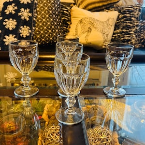 May include: A set of clear, stemmed glasses with a ribbed design. The glasses are displayed on a reflective surface, with decorative cushions in the background. The glassware is suitable for serving water, wine, or other drinks.