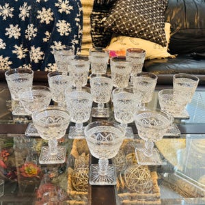 Puede incluir: Una colección de cristalería transparente tallada, que incluye copas, cuencos pequeños y vasos de varios tamaños. La cristalería presenta intrincados patrones geométricos y se asienta sobre una superficie reflectante. La disposición sugiere un ambiente formal o una estética vintage.