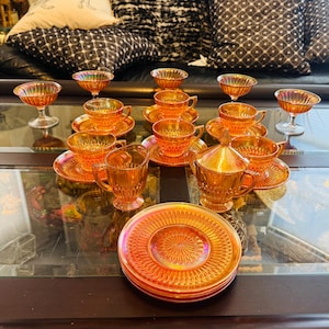 Puede incluir: Un conjunto de cristalería naranja iridiscente, que incluye tazas de té con platillos, cuencos de postre y una azucarera con tapa. La cristalería tiene un patrón texturizado y está dispuesta sobre una mesa de cristal. El conjunto es probablemente vintage.