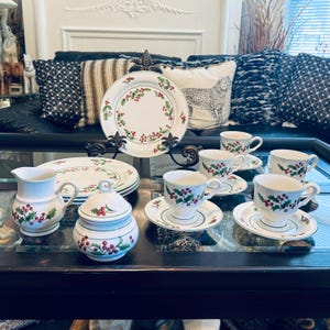 May include: A white ceramic tea set with a holly berry design. The set includes cups, saucers, plates, a creamer, and a sugar bowl. The rims and edges are trimmed with green and red accents, perfect for holiday gatherings.