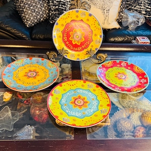Könnte beinhalten: Eine Sammlung farbenfroher dekorativer Teller mit aufwendigen Blumenmustern. Die Teller zeigen eine lebendige Farbpalette aus Gelb, Rot, Blau und Grün. Die Teller werden auf einer Glasoberfläche präsentiert, die ihre dekorativen Muster hervorhebt.