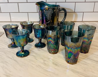 May include: A set of iridescent dark blue carnival glass featuring a pitcher and eight matching drinking vessels. Each piece displays a raised grape and leaf pattern with a metallic sheen that reflects purple, green, and gold tones.