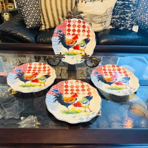 May include: A set of four decorative ceramic plates with a rooster design. Each plate has a white base with a red and white checkered pattern and a blue striped border. The rooster is depicted in bright colours.