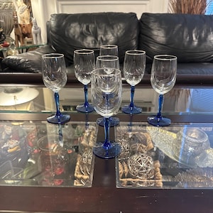 Pode incluir: Seis copos de vinho transparentes com hastes azuis e bordas douradas sobre uma mesa de madeira escura com tampo de vidro. A mesa tem itens decorativos no interior.
