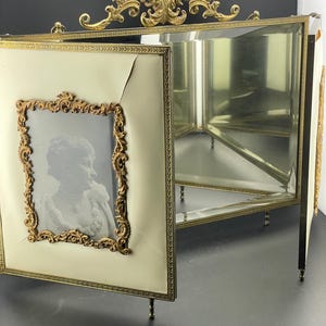 May include: An antique, three-panel photo frame with a cream-colored finish and ornate gold-colored trim. One panel displays a black and white portrait within a decorative gold frame. The frame has small feet and a decorative top.