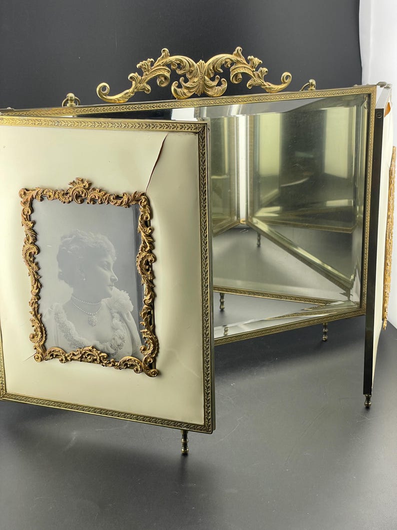 May include: An antique, three-panel photo frame with a cream-colored finish and ornate gold-colored trim. One panel displays a black and white portrait within a decorative gold frame. The frame has small feet and a decorative top.