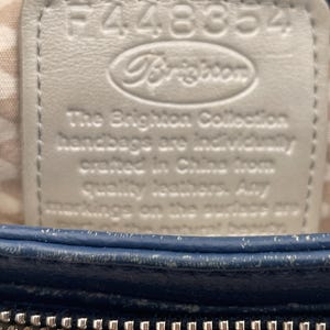 May include: Close-up of a blue leather handbag with a silver zipper. A white leather tag is embossed with the brand name "Brighton" and the text "The Brighton Collection handbags are individually crafted in China from quality leathers."