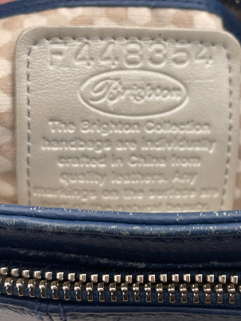 May include: Close-up of a blue leather handbag with a silver zipper. A white leather tag is embossed with the brand name "Brighton" and the text "The Brighton Collection handbags are individually crafted in China from quality leathers."
