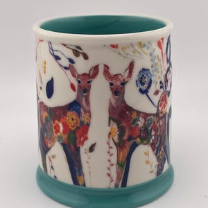 May include: A ceramic mug with a white exterior featuring two deer adorned with colorful floral patterns. The mug has a teal base and interior, with a white rim. The design includes red, blue, and yellow flowers.