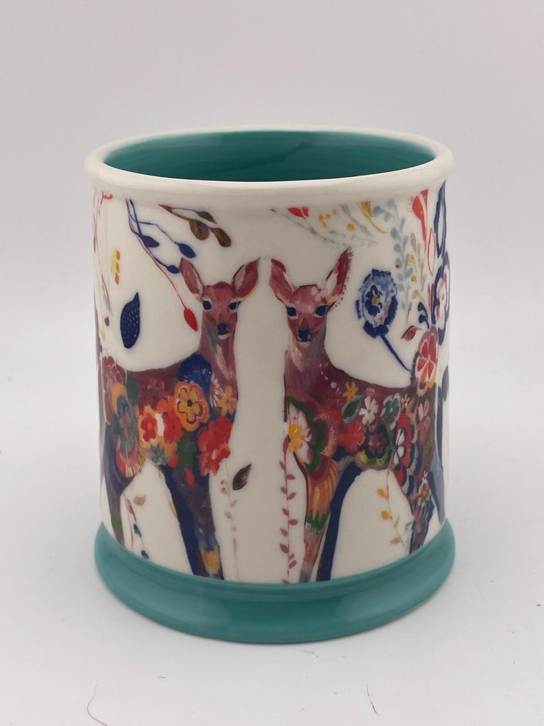 May include: A ceramic mug with a white exterior featuring two deer adorned with colorful floral patterns. The mug has a teal base and interior, with a white rim. The design includes red, blue, and yellow flowers.