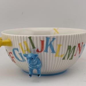 May include: White ceramic bowl with a ribbed texture, featuring colorful alphabet letters and a blue lamb figurine. The bowl has yellow handles and a blue base, perfect for serving cereal or soup.
