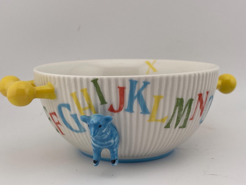 May include: White ceramic bowl with a ribbed texture, featuring colorful alphabet letters and a blue lamb figurine. The bowl has yellow handles and a blue base, perfect for serving cereal or soup.