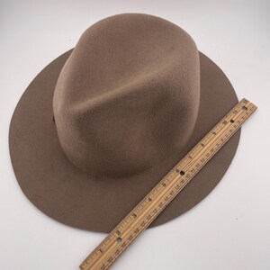 Puede incluir: Un sombrero fedora marr&oacute;n con ala ancha. Una regla de madera se coloca junto al sombrero. El sombrero parece estar hecho de fieltro o un material similar. La regla mide aproximadamente 30 cm.