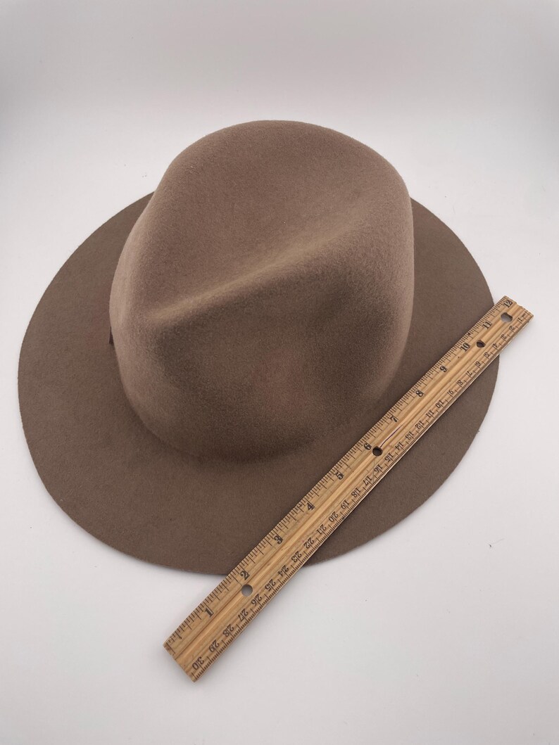 Puede incluir: Un sombrero fedora marr&oacute;n con ala ancha. Una regla de madera se coloca junto al sombrero. El sombrero parece estar hecho de fieltro o un material similar. La regla mide aproximadamente 30 cm.