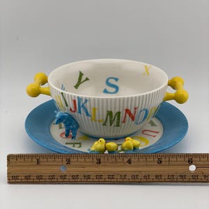 May include: A vintage children's alphabet bowl and saucer set. The white bowl has yellow handles and is decorated with colorful letters. The blue saucer features a matching alphabet design and small animal figurines. The set measures approximately 6 inches in diameter.