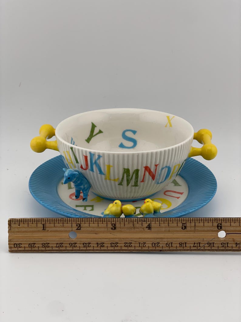 May include: A vintage children's alphabet bowl and saucer set. The white bowl has yellow handles and is decorated with colorful letters. The blue saucer features a matching alphabet design and small animal figurines. The set measures approximately 6 inches in diameter.