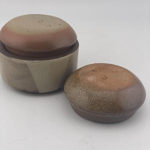 May include: Two ceramic containers with lids, featuring a matte beige and brown finish. Each piece is stamped with the number 3 and the text MADE IN FRANCE. The containers have a cylindrical base and a rounded lid with a textured, earthy appearance.