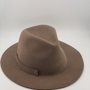 Puede incluir: Un sombrero fedora marr&oacute;n con ala ancha y una banda alrededor de la corona. El sombrero est&aacute; hecho de un material suave y tiene un dise&ntilde;o cl&aacute;sico. La banda est&aacute; asegurada con peque&ntilde;as remaches met&aacute;licos.