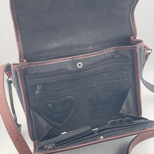 May include: Open black crossbody bag with brown leather trim and strap. The interior has multiple compartments, including a heart-shaped mirror. The bag is open, revealing the interior details.