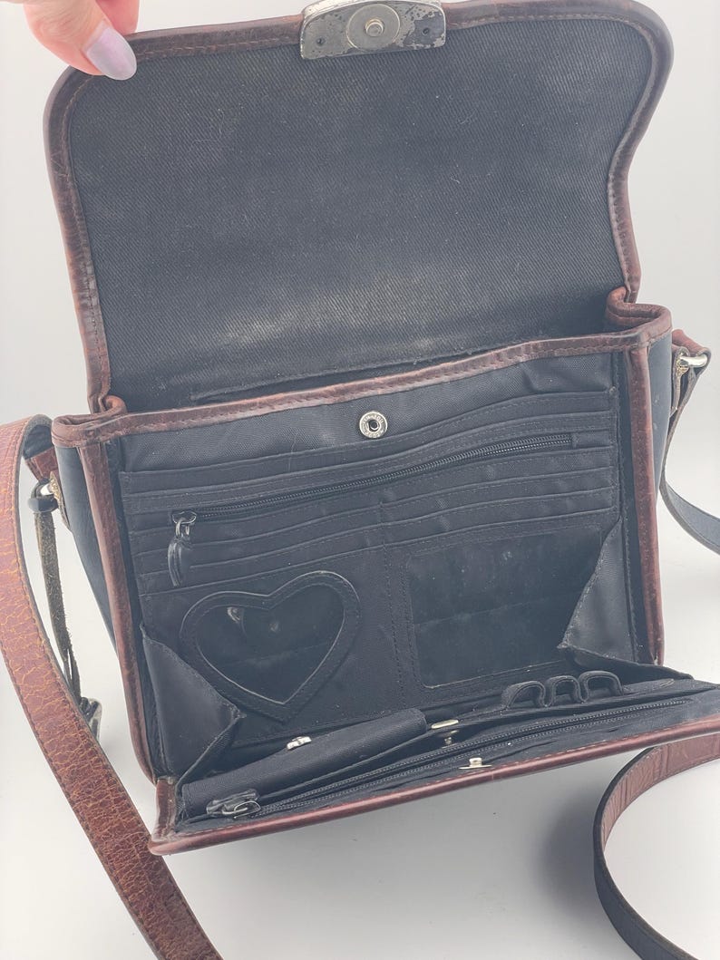May include: Open black crossbody bag with brown leather trim and strap. The interior has multiple compartments, including a heart-shaped mirror. The bag is open, revealing the interior details.