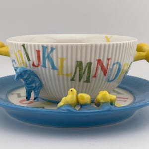 May include: A white ceramic alphabet soup bowl with a blue saucer. The bowl has yellow handles and is decorated with colorful letters and animal figurines. The saucer is blue with a matching alphabet design.