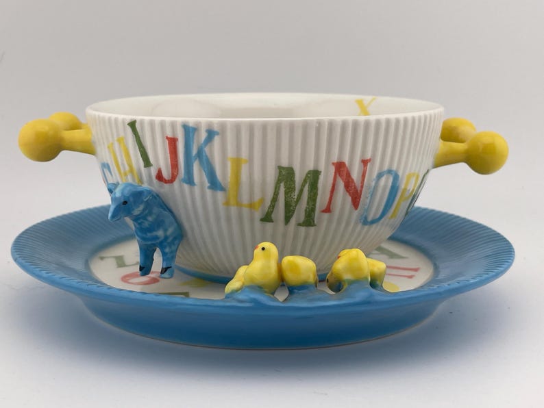 May include: A white ceramic alphabet soup bowl with a blue saucer. The bowl has yellow handles and is decorated with colorful letters and animal figurines. The saucer is blue with a matching alphabet design.