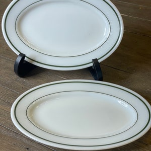 May include: Two oval-shaped white ceramic plates with a green rim. The plates are on a wooden surface.