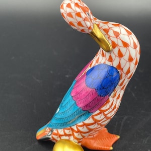 May include: A decorative porcelain duck figurine with a gold beak and feet, and a gold egg. The duck has a white body with an orange and white pattern, and colorful wings in blue, pink, and orange.