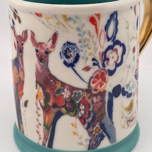 May include: White ceramic mug with a teal interior and base, featuring a colorful floral and deer design. The handle is gold. The deer are decorated with flowers in red, blue, and yellow. The mug is approximately 4 inches tall.