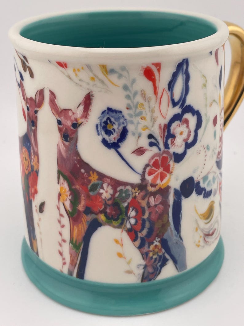 May include: White ceramic mug with a teal interior and base, featuring a colorful floral and deer design. The handle is gold. The deer are decorated with flowers in red, blue, and yellow. The mug is approximately 4 inches tall.