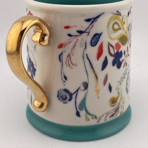 May include: A ceramic mug with a gold handle and a teal base and interior. The mug is decorated with colorful floral patterns in red, blue, yellow, and green. The mug is approximately 4 inches tall.