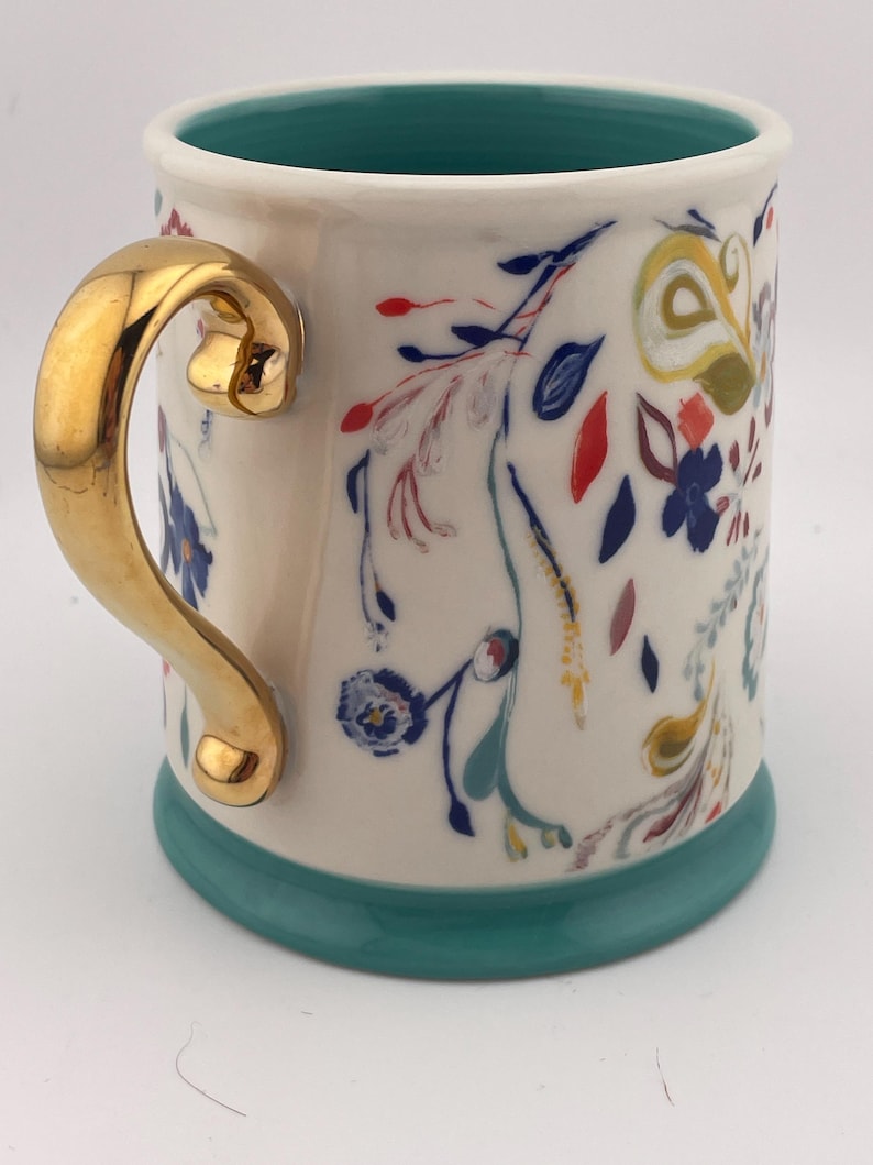 May include: A ceramic mug with a gold handle and a teal base and interior. The mug is decorated with colorful floral patterns in red, blue, yellow, and green. The mug is approximately 4 inches tall.